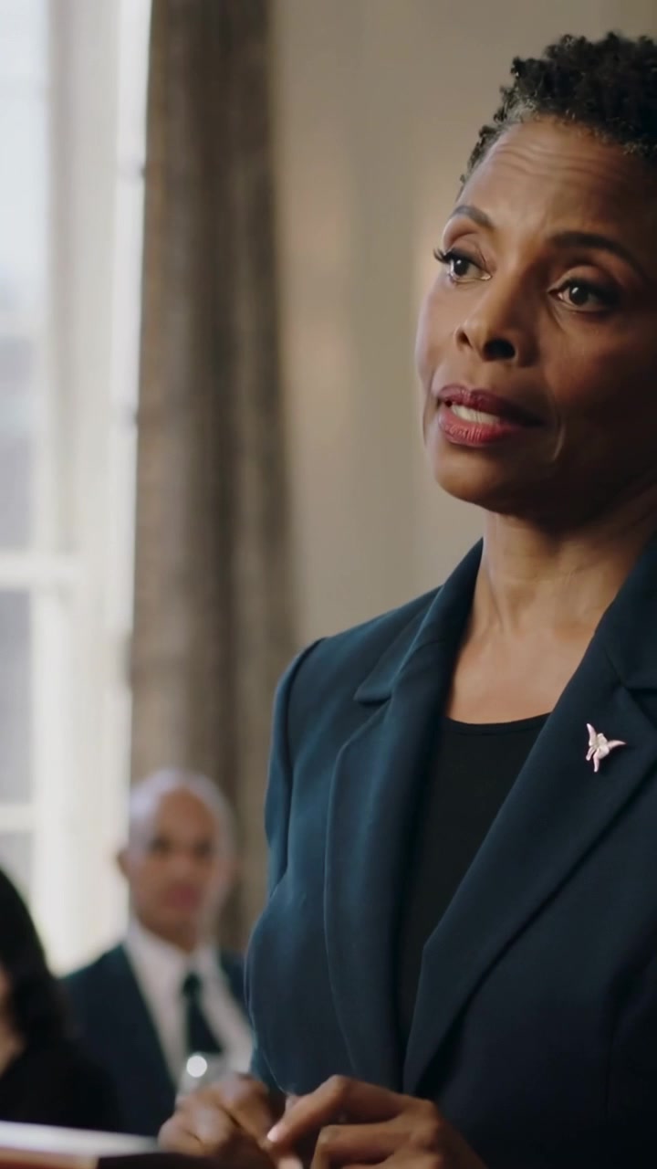 Vertical video: Taking wooden podium, woman presenting to panel at hearing, in dark blazer with pin