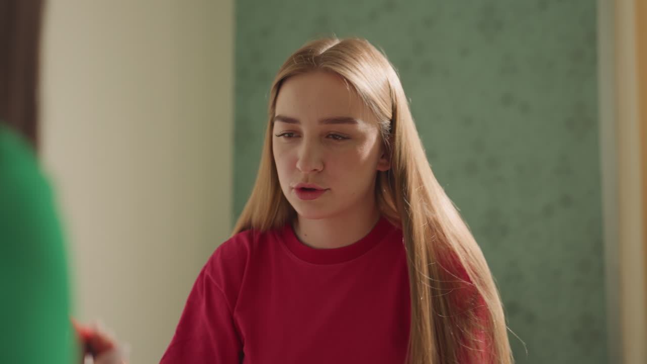 Una joven con un jersey rojo sentada en la cama, escuchando atentamente una voz fuera de cámara con expresión pensativa, pelo largo y rubio, luz natural suave, pared en tonos pastel apagados, escena doméstica íntima que transmite