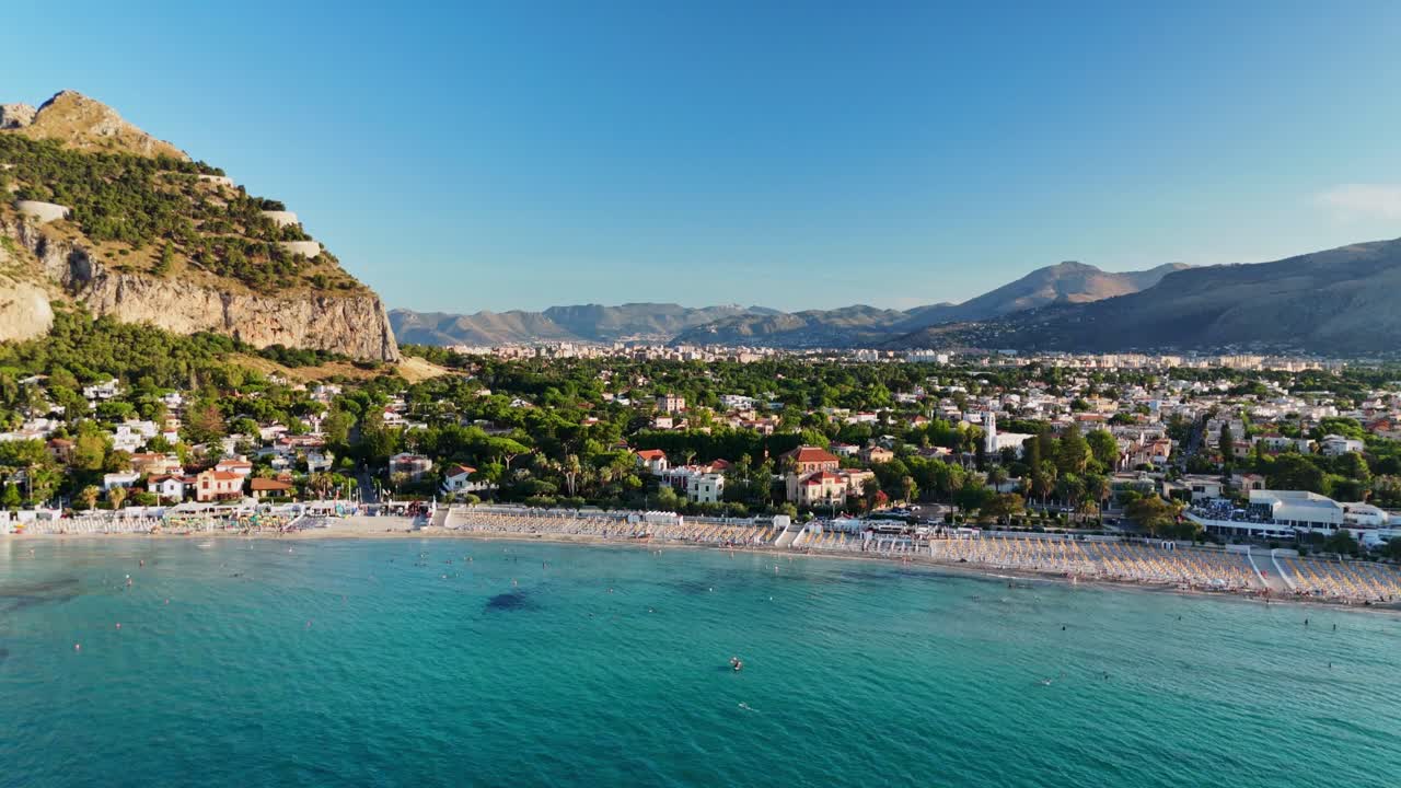 Sunny beach scene with mountains, calm sea, and vibrant houses