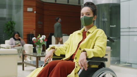 masked businesswoman in wheelchair posing for camera in office center