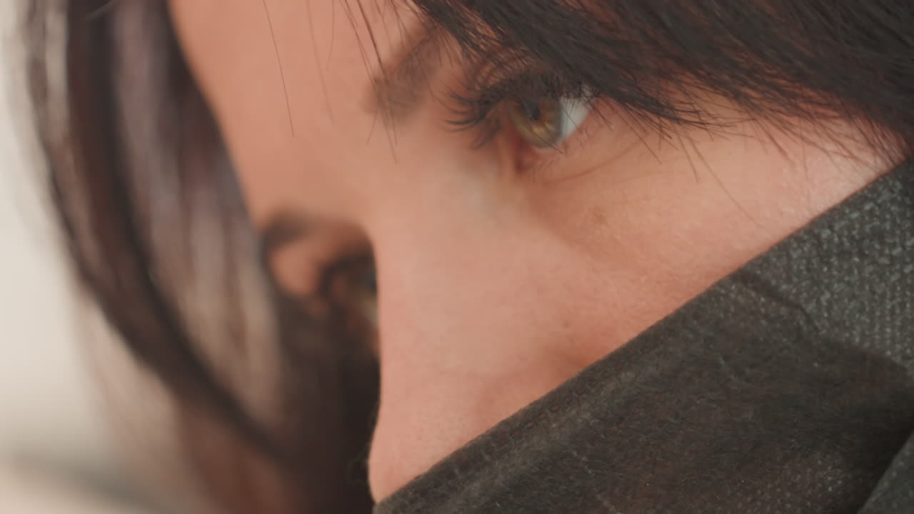 Wanita Kaukasia close-up mengenakan masker, tatapan tajam dan pencahayaan hangat lembut, rambut cokelat membingkai wajah, riasan halus, depth of field dangkal, suasana kontemplatif yang menunjukkan kerentanan, pekerja jarak jauh