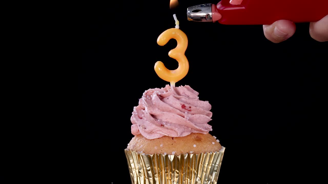 A hand uses a lighter to ignite a number three birthday candle atop a strawberry frosted cupcake, set against a black background with dramatic lighting