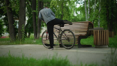 boy parks his bicycle, turning it upside down as he touches the tires and pedals, then sits on a wooden bench, the serene park is lined with trees, greenery, benches and residential building