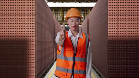 asian female engineer with safety helmet disapproving with no index finger sign while standing with red brick packed in stacks are stored