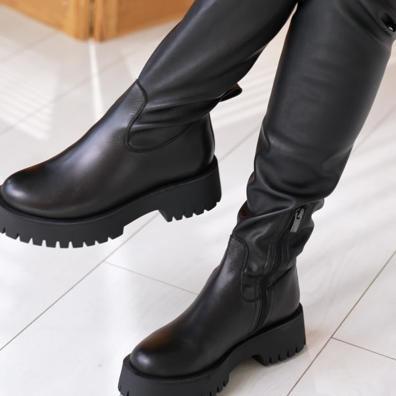 New eco leather boots on massive plain soles. Model demonstrates black autumn footwear sitting in studio. White floor backdrop
