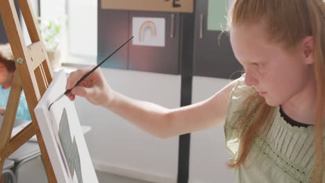 video of happy caucasian schoolgirl concentrating at easel, painting in art class, copy space