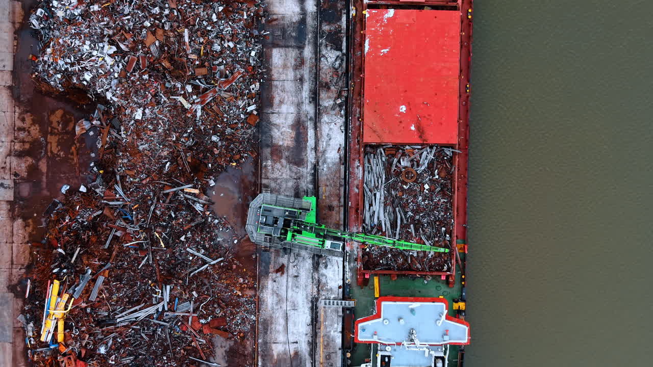 Gru industriale che carica rottami metallici dal molo alla nave. Veduta dall'alto di una gru verde che sposta rifiuti metallici arrugginiti da un grande mucchio sul molo nella stiva della nave