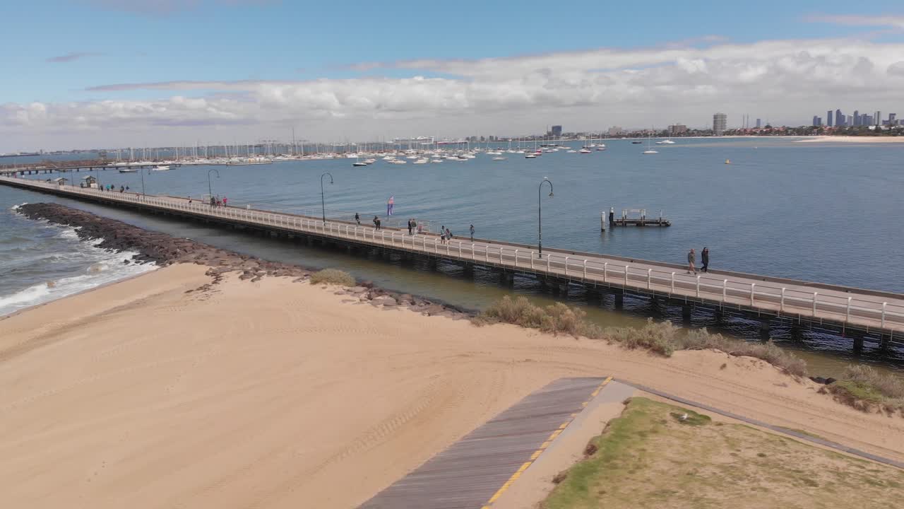 태양이 비치는 동안 모래 해변, 바닷물, 멜버른 스카이라인을 배경으로 세인트 킬다 해변의 해안을 보여주는 공중 궤도 샷