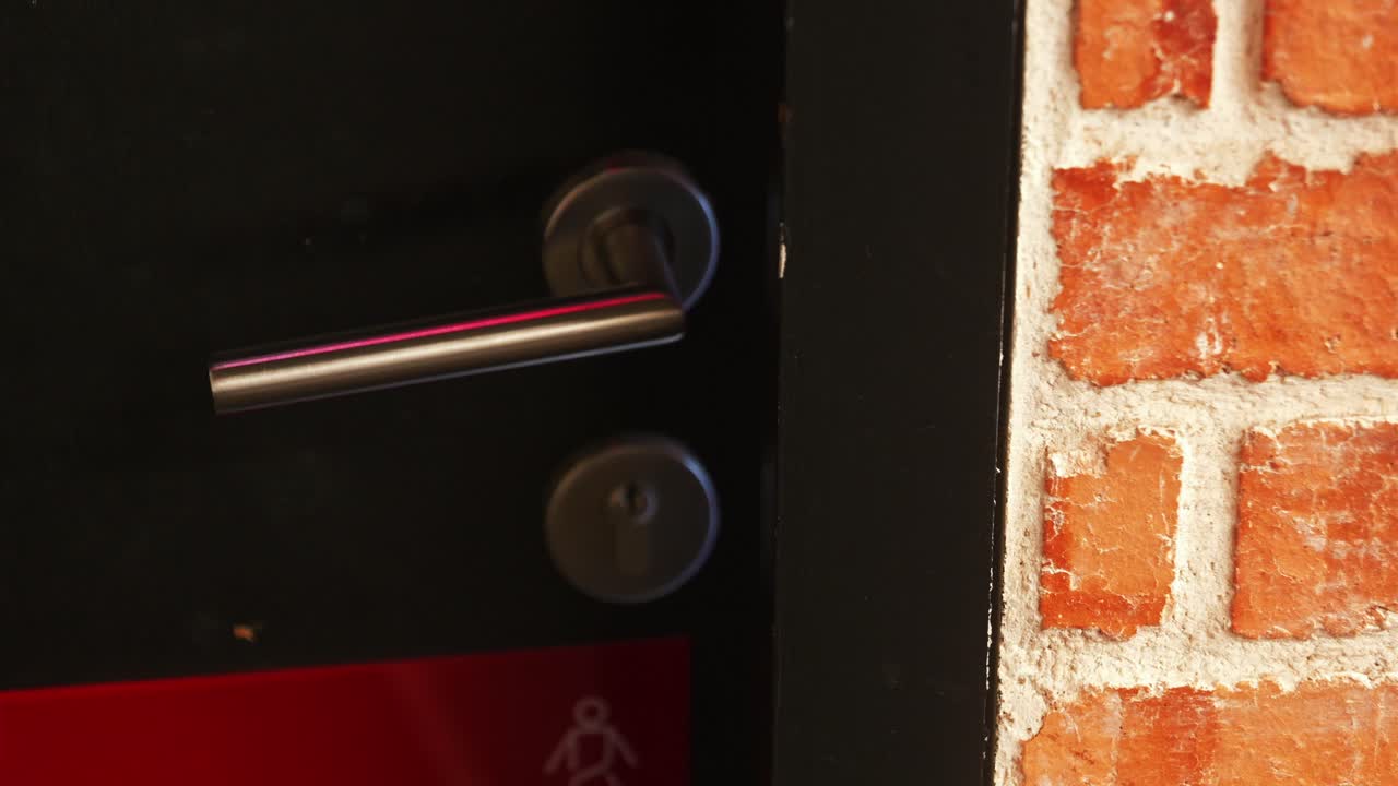 A close-up of a silver metal lever handle being pushed down to open a dark fire door, adjacent to an exposed red brick wall, emphasizing motion and contrast