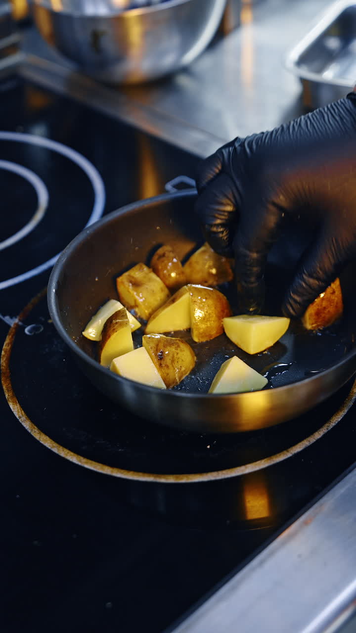 Large pieces of potato in peels are fried in oil. Gloved hand turns the pieces on the frying pan. Close up. Vertical video.