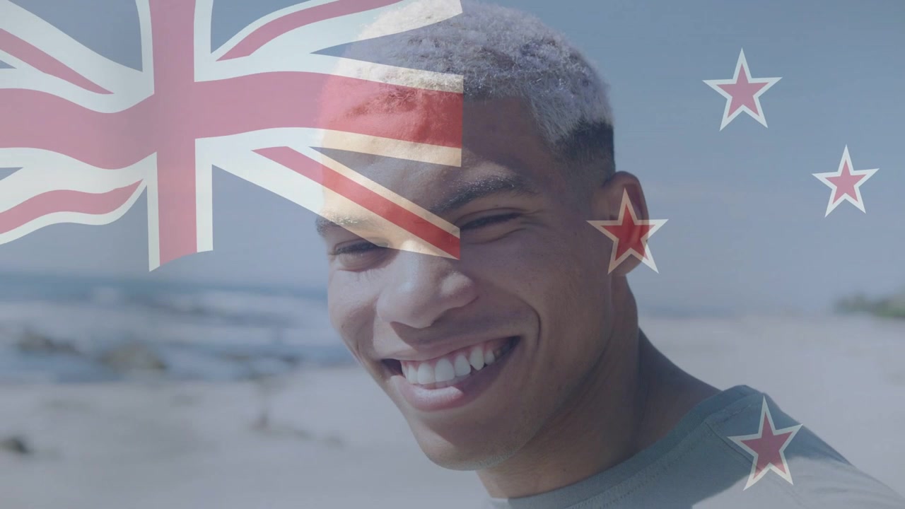 Man draait hoofd naar camera op het strand, merkt een vlag-overlay op, glimlach wordt breder ter promotie van Nieuw-Zeelands toerisme.