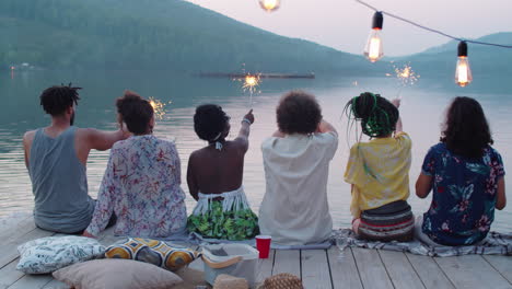 friends sitting on lake pier and dancing with sparklers