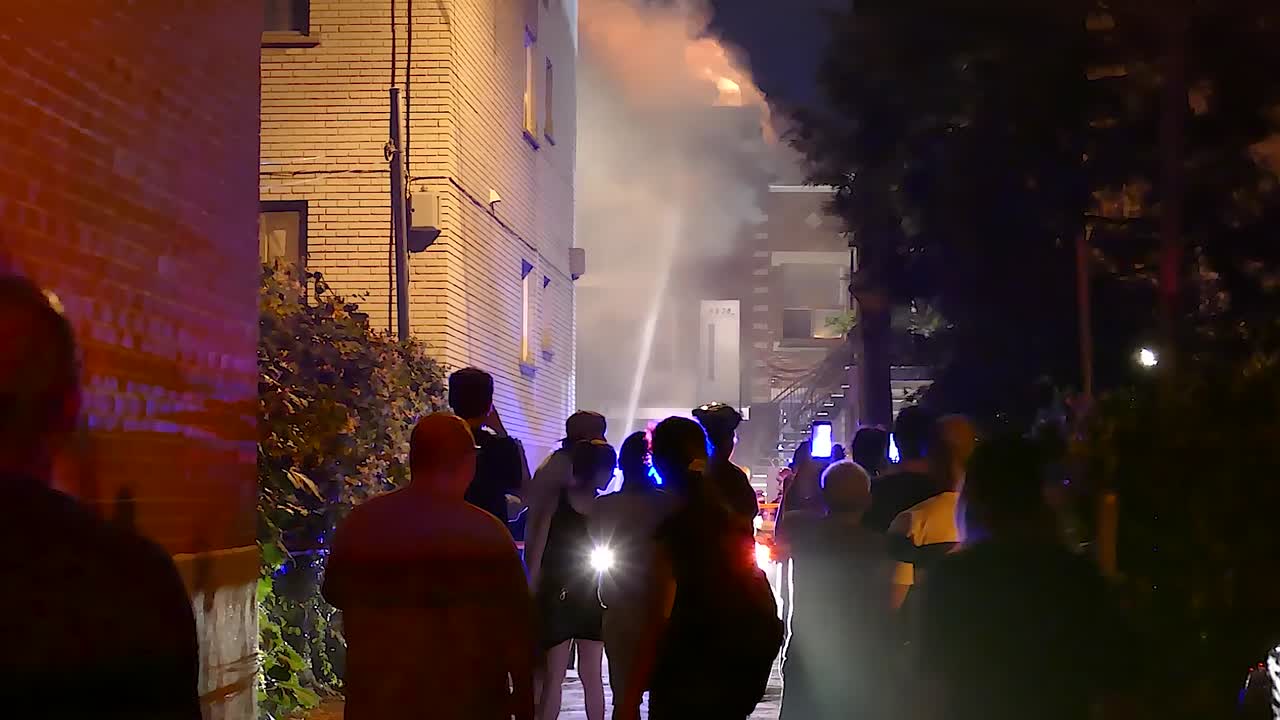 Firefighters battle building fire as people record scene on their phones in downtown Montreal, Quebec at night.