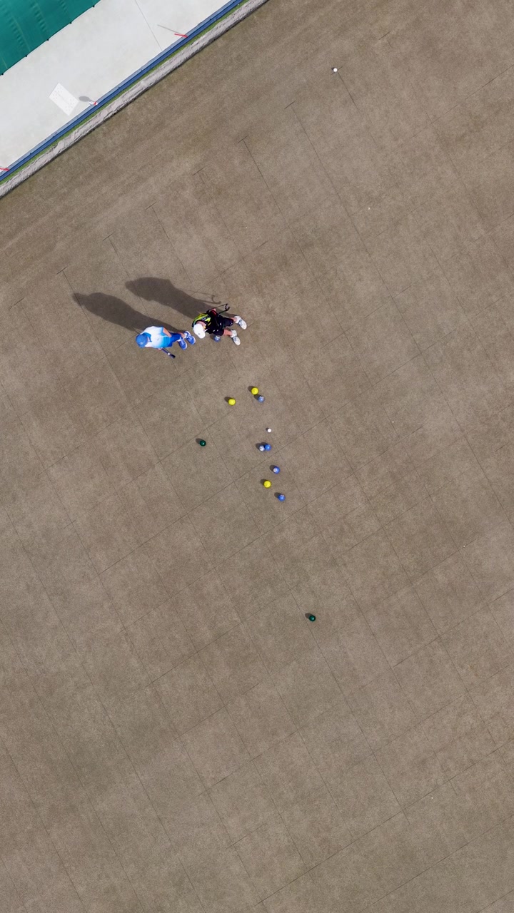Ripresa aerea con drone di giocatori che lanciano bocce verso il boccino su un campo verde all'aperto e soleggiato.