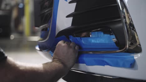 mechanic using paper tape to mask front bumper of car to prepare for painting