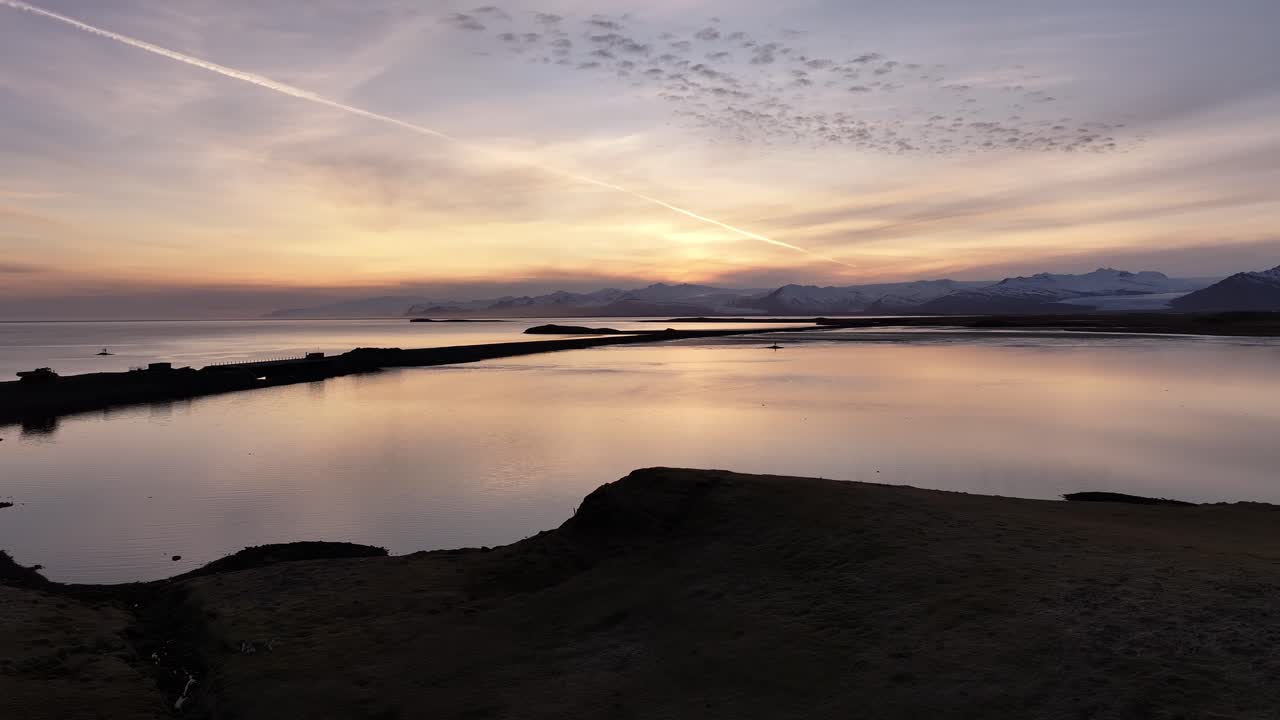 Aerial drone shot of a serene coastal lagoon at sunset near Höfn, Iceland, with calm waters reflecting the golden sky and distant snow-covered mountains.