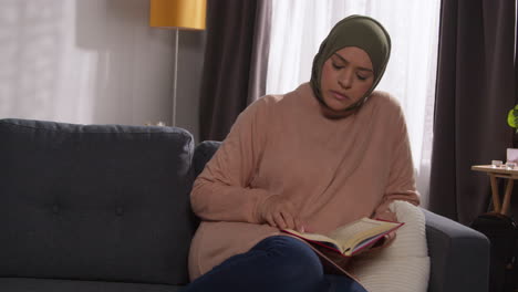 muslim woman wearing hijab sitting on sofa at home reading or studying the quran 2