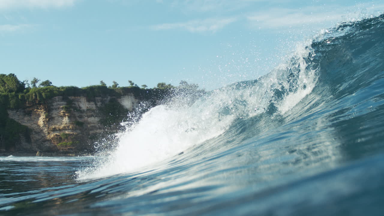 Tropical ocean wave breaks near dark rocky coastline in vibrant light