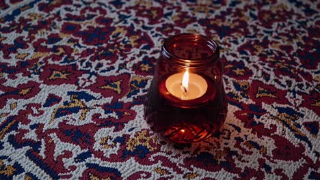 small round candle is burning brightly in a red glass candle holder placed on an ornate tablecloth, creating a warm and inviting atmosphere