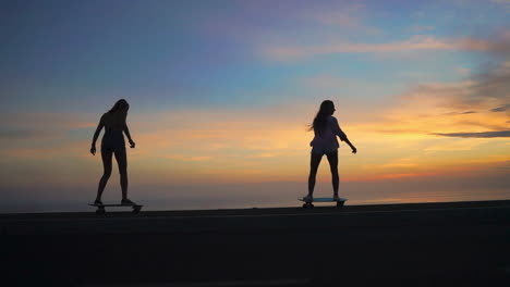 As-daylight-wanes,-two-friends-ride-skateboards-on-a-road,-the-moment-slowed-down-to-a-mesmerizing-pace.-Mountains-and-a-lovely-sky-complete-the-backdrop,-while-they-sport-shorts