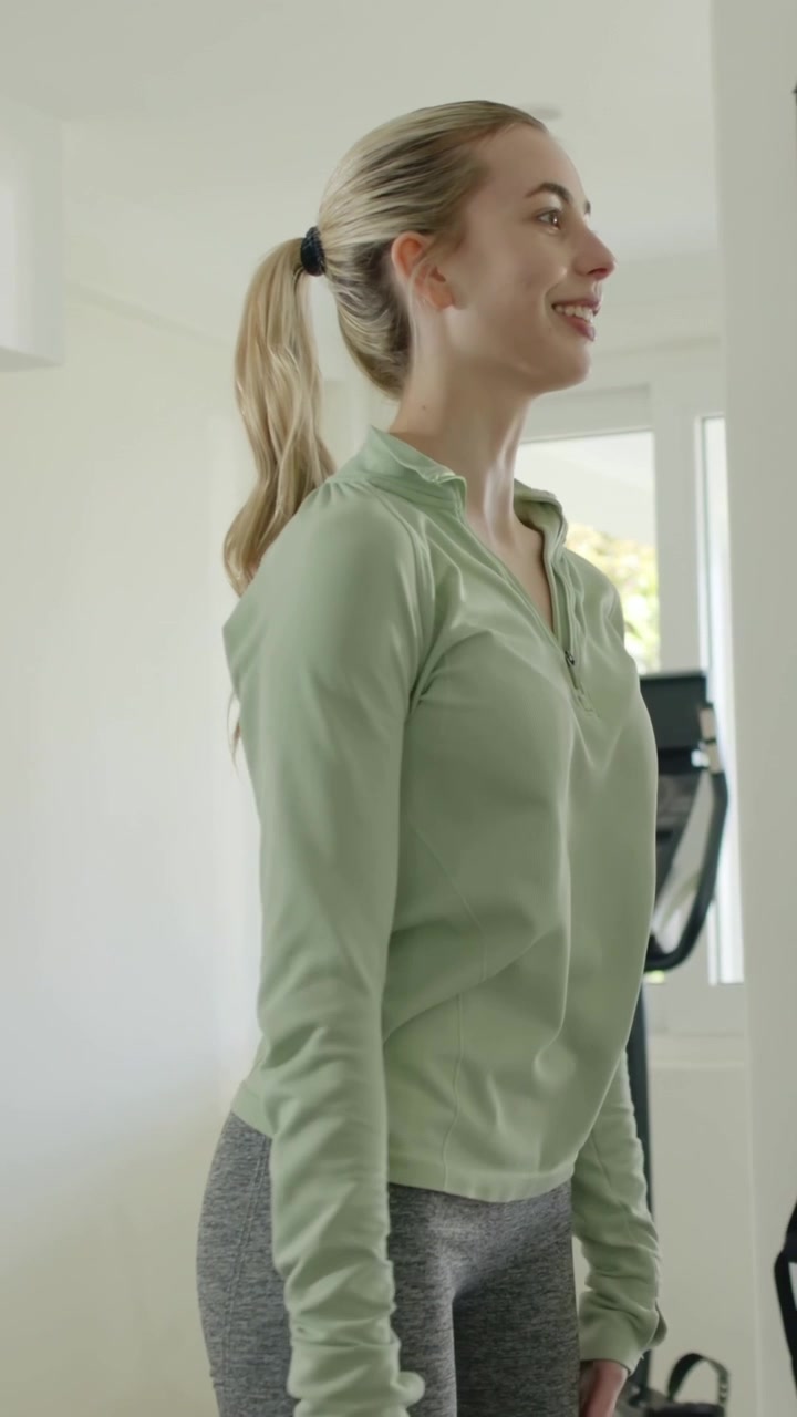 Vertical video: Starting hands on hips, woman doing shoulder warmup in room by machine, wearing top
