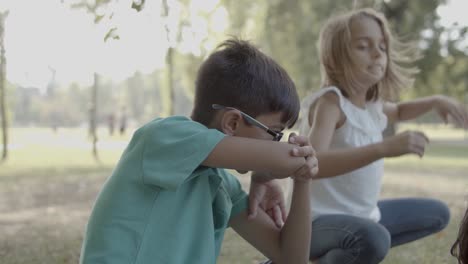 chico lindo con anteojos frotándose la nariz con el codo y hablando con una chica mientras se sientan en la hierba en el parque