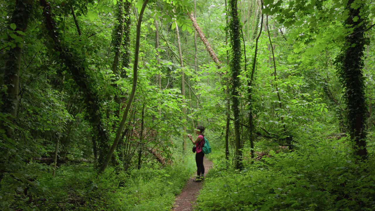 Wide slow-motion of a female hiker walking along a narrow forest path, pausing to gently tap the leaves.
