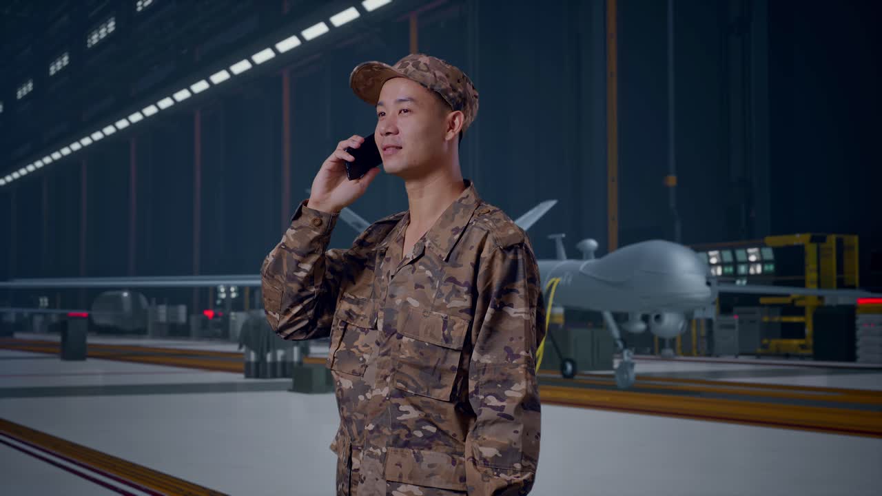 Soldato uomo asiatico di profilo che parla al cellulare e si guarda intorno mentre si trova in piedi con un moderno drone militare in un hangar di manutenzione