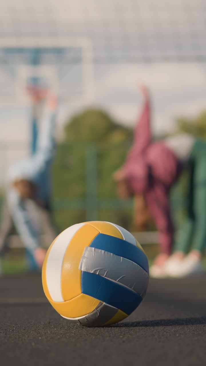 야외 코트에서 휴식을 취하는 배구 선수의 클로즈업, 배경에는 스트레칭과 휘어짐을 수행하는 운동 선수의 흐릿한 시야, 스포츠 시설 및 건물이 보입니다.