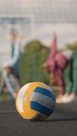 primer plano de una pelota de voleibol descansando en una cancha al aire libre con vista borrosa de los atletas realizando estiramientos y curvas en el fondo, instalación deportiva y edificio visible