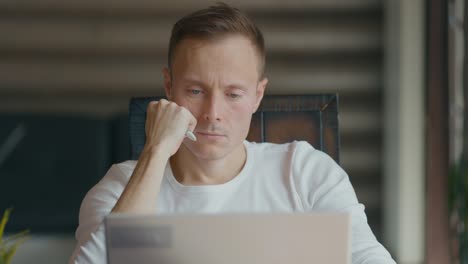 concentrated young man sits at table and looks into laptop. holding right hand to face. close-up