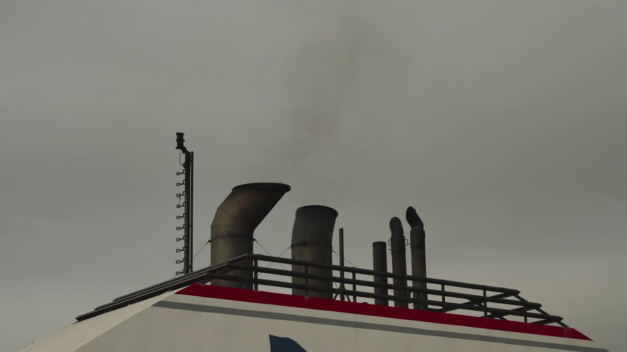 Multiple exhaust funnels and antenna mast rise from ferry upper deck with red stripe under grey sky