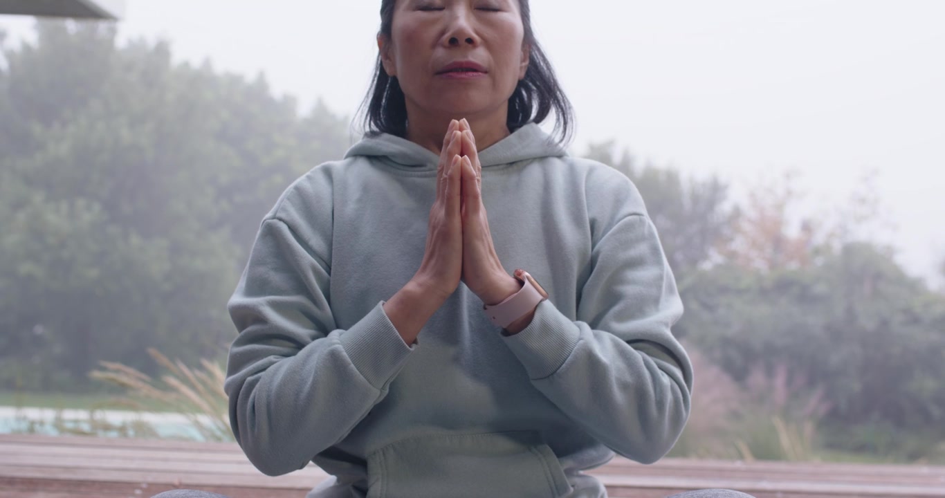 Settling on blue mat, Asian woman in hoodie and leggings raising palms, meditating on wooden deck