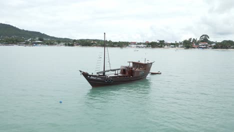 Aerial-view-circling-large-Thai-fishing-boat-anchored-offshore-with-dingy-attached