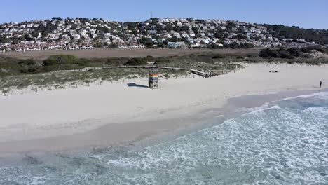 Drone-flying-backwards-reveals-surf-club-with-waves-washing-against-the-shoreline-in-Son-Bou-beach,-Menorca