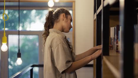 Chica-Estudiante-Eligiendo-Un-Libro-En-La-Biblioteca-En-Una-Estantería