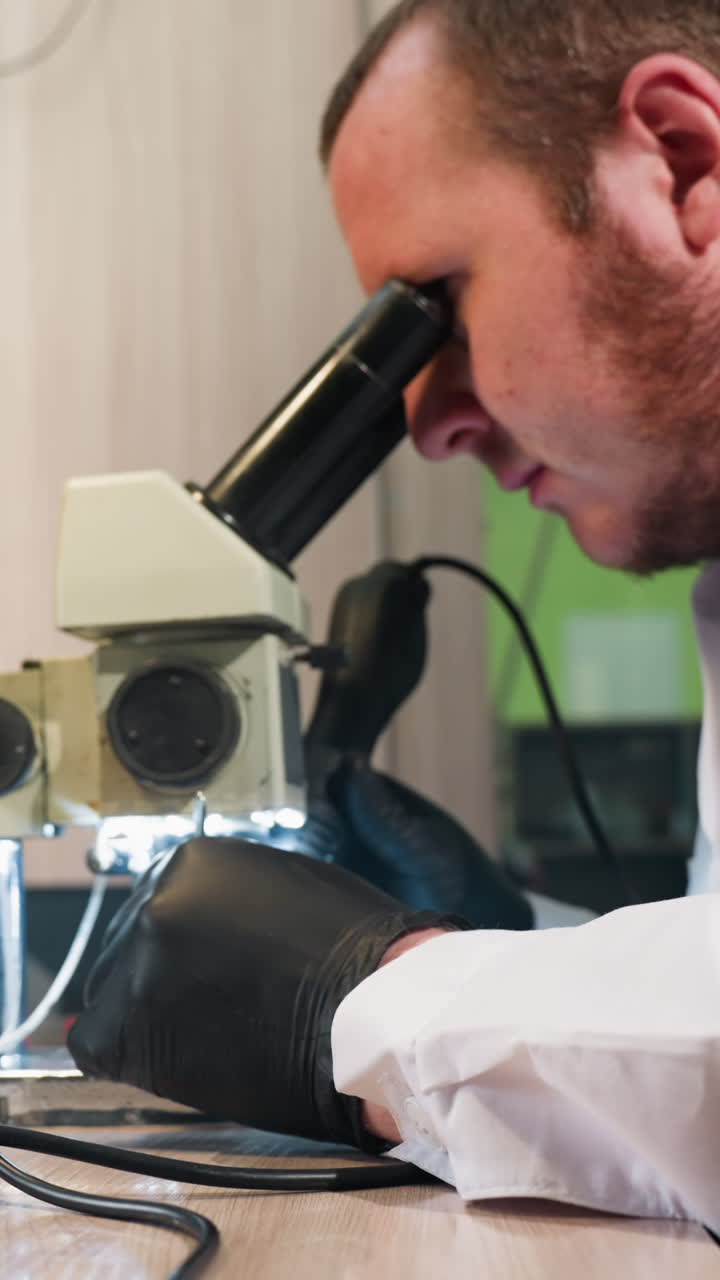 Close view of a technician in a white lab coat using a microscope having light to work on circuit board, the workbench is filled with various tools
