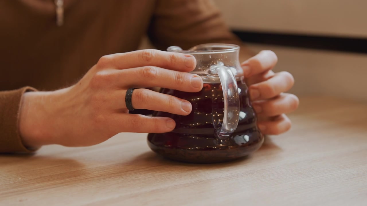 Primo piano delle mani di un uomo che tengono e muovono leggermente una caraffa di caffè in vetro riempita con liquido scuro su un tavolo di legno. La scena presenta un'illuminazione soffusa, un'espressione calma e dettagli come un anello nero indossato.