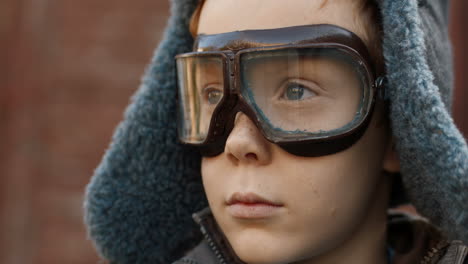 close up of the face of little red haired boy in aviator hat and goggles smiling cheerfully at the camera