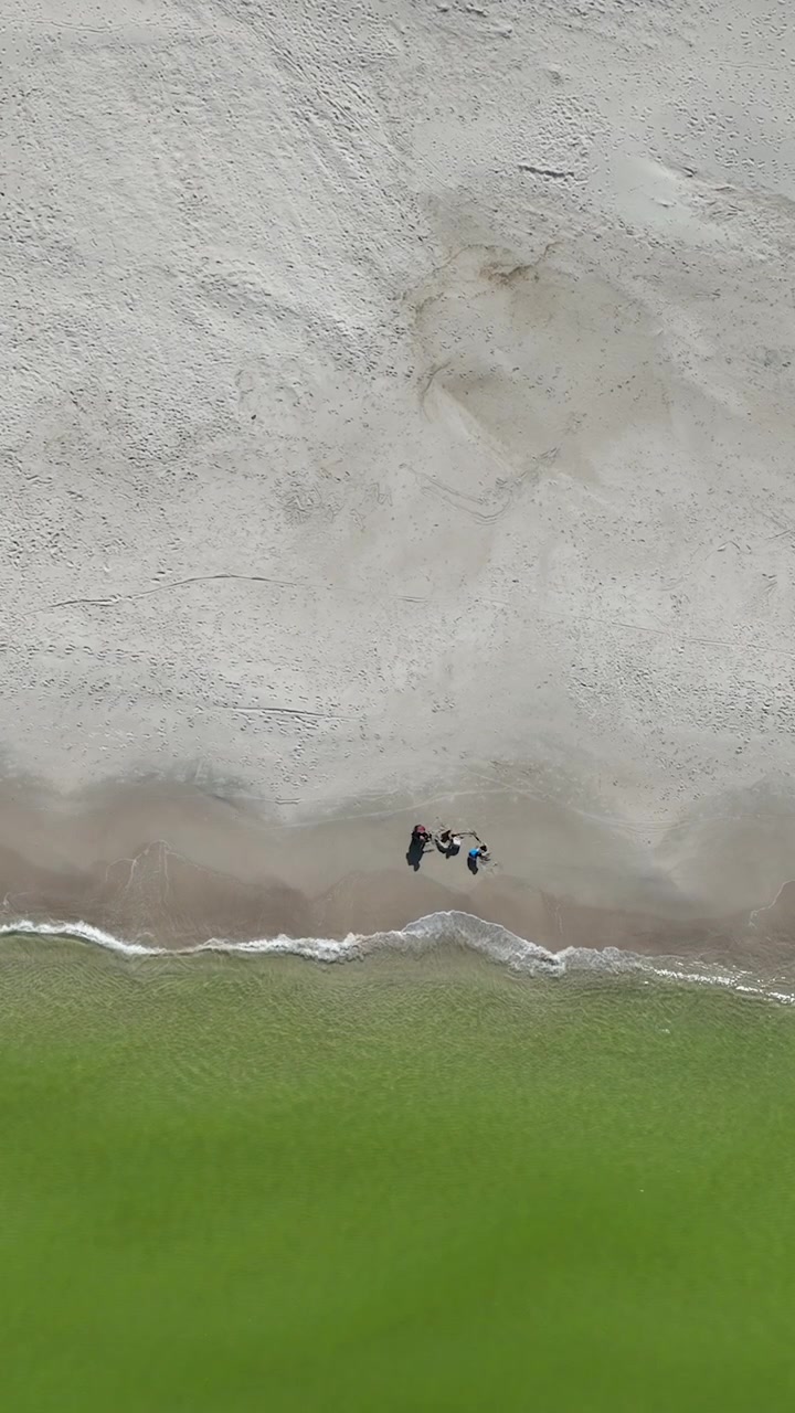 Vertikalt bilde tatt ovenfra av mennesker som går langs strandlinjen på en hvit sandstrand med grønt havvann i Osetnik, Polen. Ideelt for innhold om reise og sommerferie
