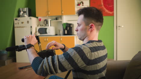 man adjusting tripod-mounted camera with microphone in home studio, focused on camera setup. comfortable indoor environment with green walls and furniture. preparing for video production work
