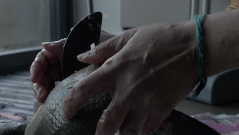 a close up shot of a lady preparing a rohu fish using a traditional bengali bonti, her hands battling to grip the slimy fish as she slices off the pelvic fin cleaning the fish before cooking