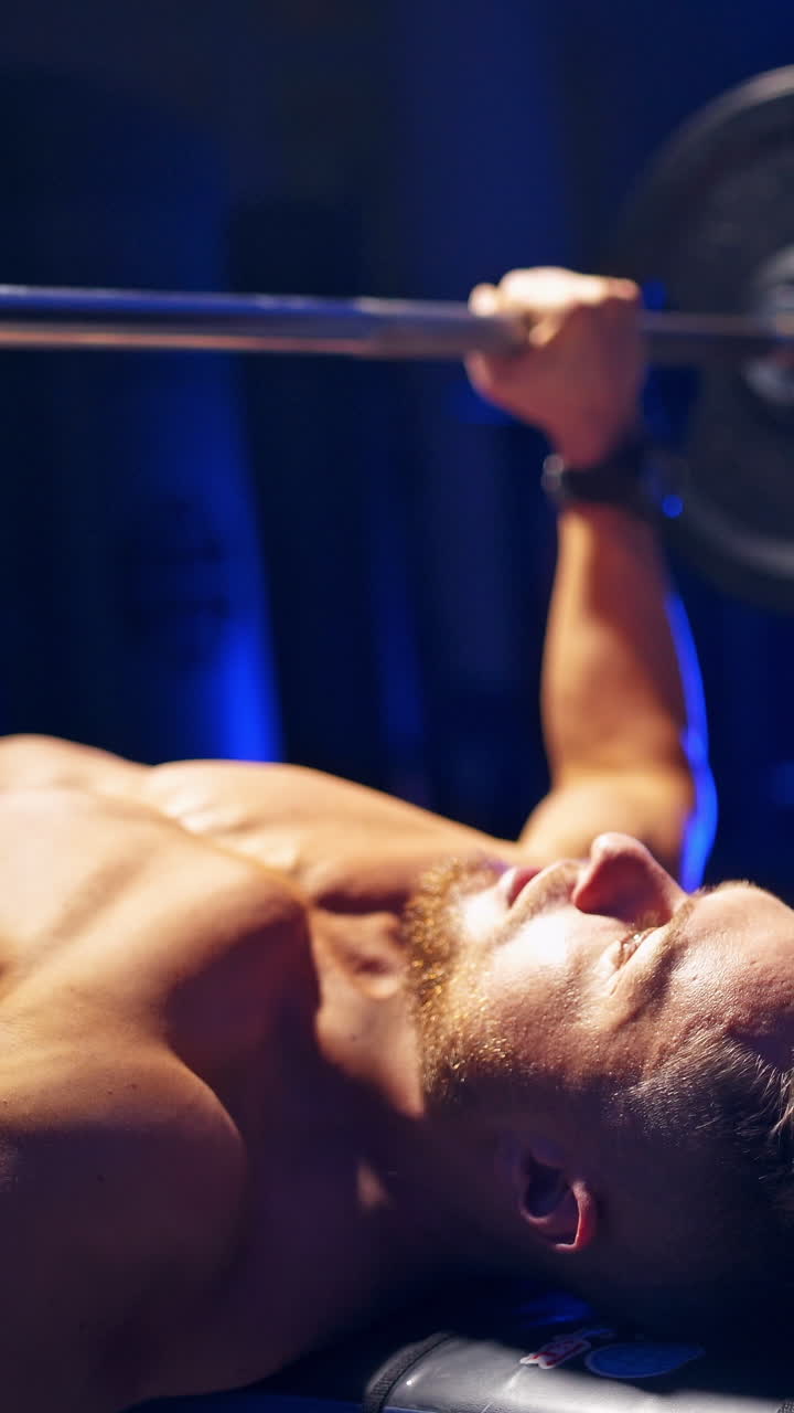 Shirtless young bodybuilder doing bench press for chest. Strong abs. Blue light filter. Video from the side. Vertical video