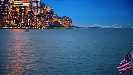 american flag waves at the front part of the boat. large white riverboat moves by the river in front. luminous skyline of new york at backdrop