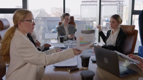 A-group-of-businesswomen-in-business-clothes-are-communicating-during-a-meeting-and-passing-each-other-sheets-with-graphs-and-ideas-about-their-future-work-in-a-modern-society-with-large-windows