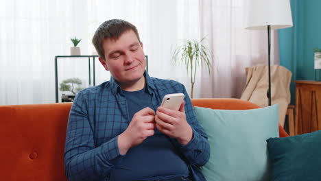 young caucasian man text messaging on smartphone sitting on sofa watching online movie at home room