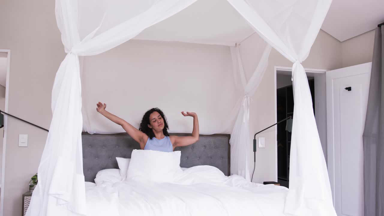 Donna africana che si sveglia, cambia posizione e allunga le braccia su un letto a baldacchino con biancheria da letto bianca.