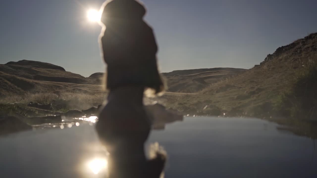 Warm and inviting shot captures a woman, silhouetted against a bright, low sun, enjoying the tranquil waters of Hrunalaug hot spring in Iceland