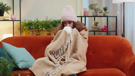 sick teen girl kid wear hat wrapped in plaid sit alone shivering from cold on sofa drinking hot tea