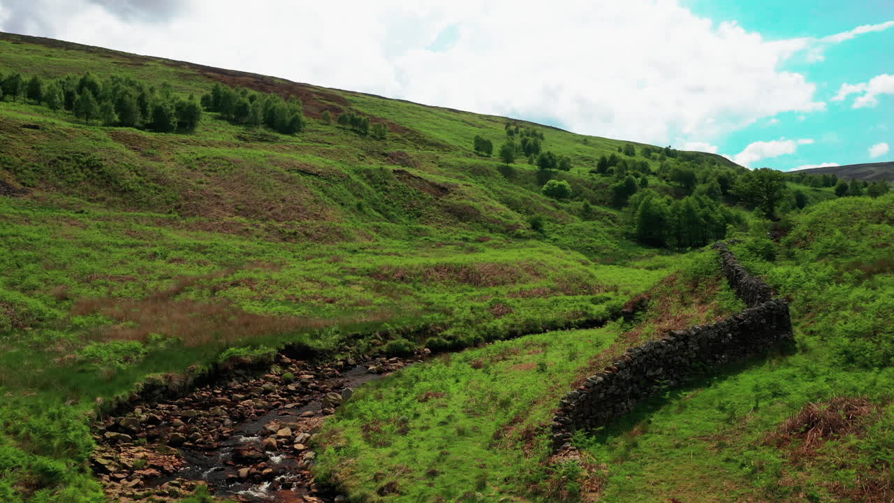 toma aérea ascendente con vistas a un arroyo rocoso, rodeado de colinas verdes, día soleado y brillante
